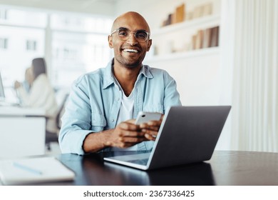 Business man smiling at the camera while holding a mobile phone. Young male designer sitting in an office with a laptop.