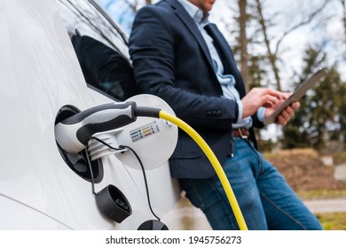 Business man standing near charging electric car or EV car and using tablet in the street. 