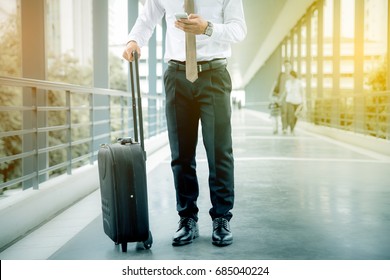 A Business Man Standing On Walkway With Wheels Luggage. His Left Hand Hold A Smart Phone And Slide Touch Screen. Shallow Depth Of Field. Warm Light.