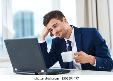 A business man in a suit looks at a laptop and holds a cup of coffee in his hand in the office - Shutterstock ID 1502394497