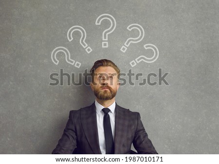 Business man thinking of answers to lots of questions. Confused bearded young man in suit sitting against background of grey concrete wall looking up at several question mark doodles above his head