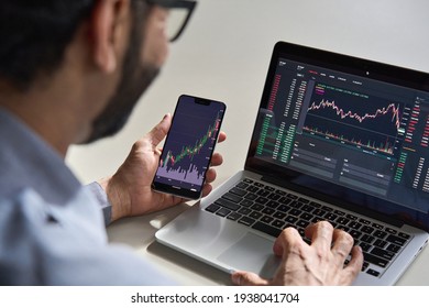 Business man trader investor analyst using mobile phone app analytics for cryptocurrency financial market analysis, trading data index chart graph on smartphone and laptop screen. Over shoulder view