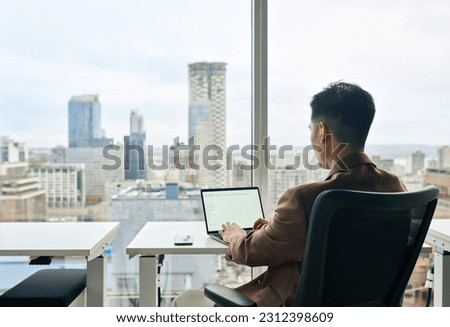 Business man using laptop in modern office city view sitting at desk. Businessman working computer with white blank mock up screen display for business websites or services ads. Over shoulder view.
