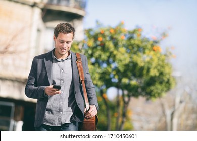 Business Man Using Mobile Phone Walking In City Street Commuting To Work With Blazer And Messenger Bag Texting On Smartphone. Young Businessman Urban Lifestyle.