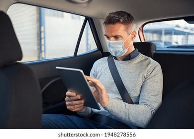 Business man wearing a face mask for safety against covid19 sitting on back seat of a car and using digital tablet. Successful entrepreneur working on digital tablet while traveling in taxi. - Powered by Shutterstock