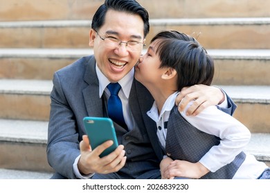 A Business Man Wearing A Suit. He Was Looking At The Mobile Phone In His Hand And Smiled Happily While His Lovely Son Was Kissing His Cheek - Dad And Son Happy Asian Family Concept