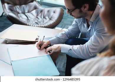 Business Man At Work In Corporate Office. Busy Hispanic Manager With Eyeglasses Working And Meeting With Young Assistant. People Sitting In Waiting Room And Signing Documents.