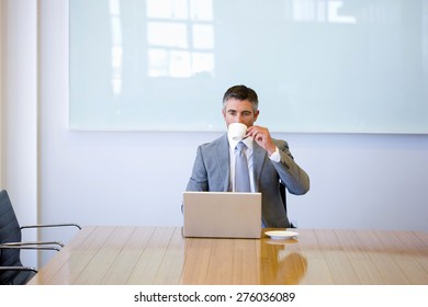 Business Manager Sitting At The End Of A Meeting Table, Drinking Cafe In Front Of His Laptop. 