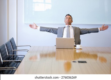 Business Manager Sitting At The End Of A Meeting Table, With Cup Of Cafe And Laptop, With Arms Outspread. 