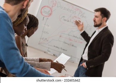 Business Meeting At Office. Handsome Man Presenting Charts On Whiteboard To Team. Multi Ethnic Group Of People