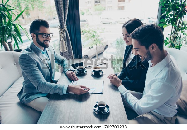 Business meeting\
at the restaurant. Happy couple is buying new house, young bearded\
attractive brunet lawyer in a formal wear is presenting contract to\
sign, they are having\
coffee