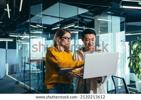 Business meeting of two employees, Asian and female colleagues, discussing the project, working in a modern office, looking at a laptop screen, discussing edits, and project success
