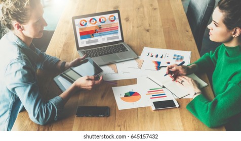 Business meeting.Two young businesswomen sitting in meeting room at table and discussing.First girl using tablet PC,second takes notes in notepad.On table laptop with graphs,charts,diagrams on screen. - Powered by Shutterstock