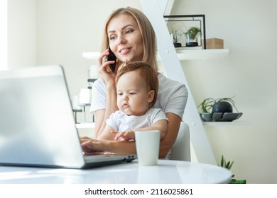 Business Mom With Little Daughter In Her Arms Uses A Laptop And Talking On Phone While Sitting At A Table At Home