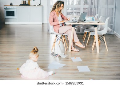 Business Mom Working With Laptop At Home While Her Daughter Drawing On The Floor. Business, Motherhood, Multitasking And Family Concept.