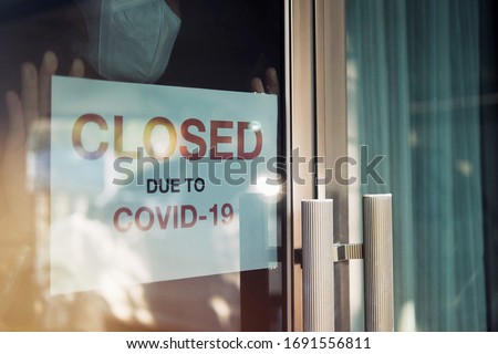 Business office or store shop is closed, bankrupt business due to the effect of novel Coronavirus (COVID-19) pandemic. Unidentified person wearing mask hanging closed sign in background on front door.