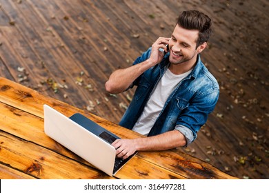 Business on fresh air. Top view of happy young man working on laptop and talking on the mobile phonewhile sitting at the wooden table outdoors - Powered by Shutterstock