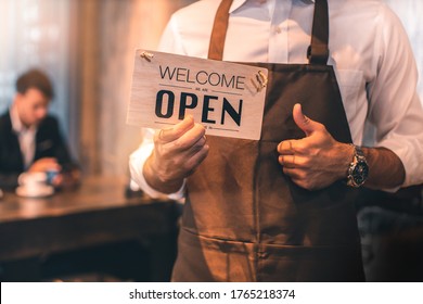 Business Owner Attractive Young Asian Man In Apron Hanging We're Open Sign On Front Door  Welcoming Clients To New Cafe. Happy Waiter With Protective Face Mask Holding Open Sign While Stand At Cafe .