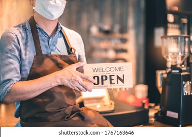 Business Owner Attractive Young Asian Man In Apron Hanging We're Open Sign On Front Door  Welcoming Clients To New Cafe. Happy Waiter With Protective Face Mask Holding Open Sign While Stand At Cafe .