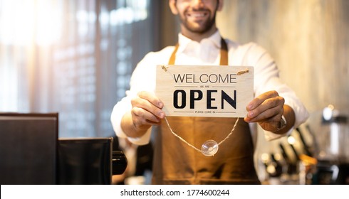 Business Owner Attractive Young Asian Man In Apron Hanging We're Open Sign On Front Door  Welcoming Clients To New Cafe. Happy Waiter With Protective Face Mask Holding Open Sign While Stand At Cafe .