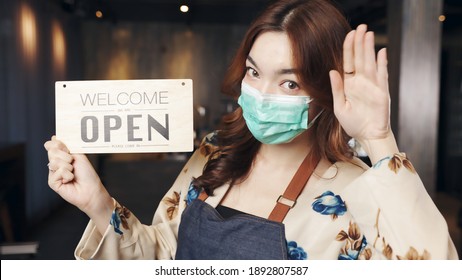 Business Owner Attractive Young Asian Woman In Apron Hanging We're Open Sign On Front Door  Welcoming Clients To New Cafe. Happy Waitess With Protective Face Mask Holding Open Sign At Cafe .