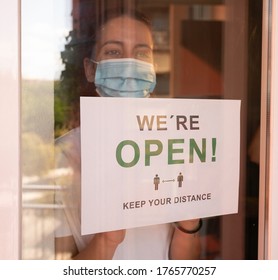 Business Owner With A Face Mask Hanging The Open Sign In Her Restaurant. Woman With A Face Mask Hanging The Open Sign In Her Store. New Normal. We Are Open. Keep Your Distance