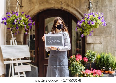 Business Owner Hispanic Woman Wear Protective Face Mask Holding Open Sign At Her Floral Shop Outdoor, Open Again After Lock Down Due To Outbreak Of Coronavirus Covid-19. Supporting Local Business.