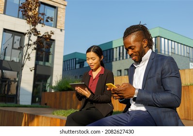 Business Partners African American Man In Navy Suit And Young Asian Brunette Woman Work With Phone And Tablet Sitting Near Office At Break