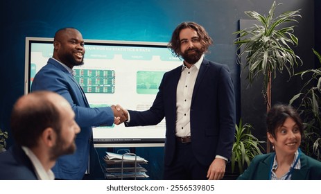 Business partners celebrate the beginning of a new project between their companies, shaking hands in a meeting. Leaders forging an agreement to develop the corporation and collect resources. Camera B. - Powered by Shutterstock