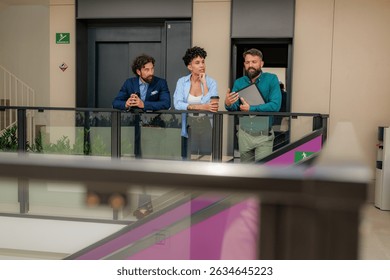 Business partners engaging in conversation during a coffee break, standing in the hallway of a modern office building, positioned near the elevators, surrounded by sleek architecture