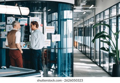 Business partners standing at infographic board discussing strategy, focusing on charts and progress tracking in open office hallway with natural light