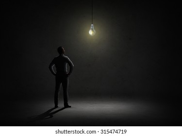 Business, People, Inspiration And Idea Concept - Businessman Looking At Lighting Bulb In Dark Room From Back