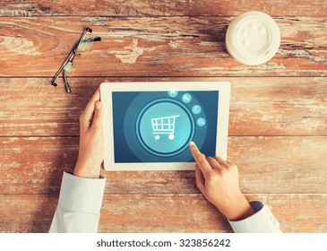 business, people, online shopping and technology concept - close up of hands pointing finger to tablet pc computer screen with trolley icon, coffee cup and eyeglasses on table