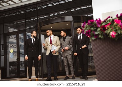 Business People Outdoor Meeting. A Group Of Male Businessmen In Suits Exit The Front Door Of A Glass Building. Working Break. Teamwork And Brainstorming. Successful Teamwork