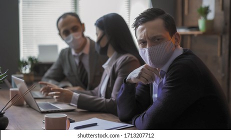 Business People In Protective Masks At Meeting In Office Despite Quarantine, Focus On Serious Young Man Communicates With Colleagues Sitting At Table In Board Room