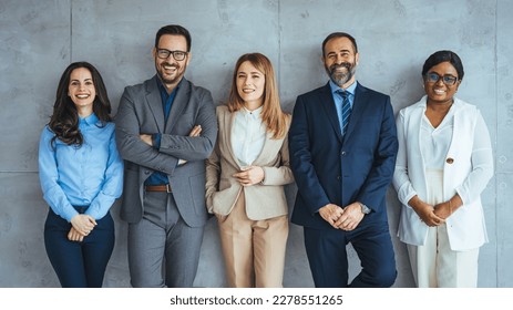 The business people standing on the gray wall background. Business team headed with boss, posing to camera over grey wall in office. Diverse businesspeople smiling at the camera  - Powered by Shutterstock