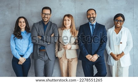 The business people standing on the gray wall background. Business team headed with boss, posing to camera over grey wall in office. Diverse businesspeople smiling at the camera 