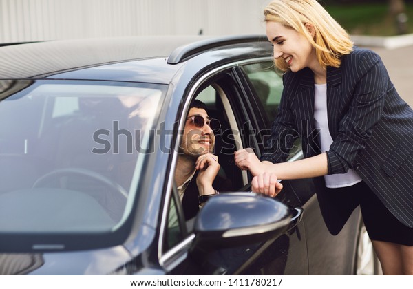 Business people\
talking near car park. The man in the glasses is sitting in the\
car, the woman stands next to\
him