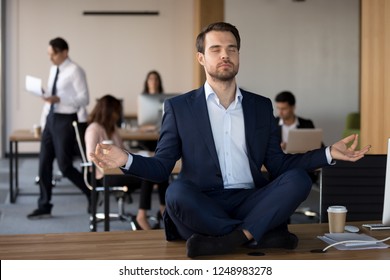Business People working in coworking space, focus on millennial peaceful employee in formal suit sitting without shoes in lotus position on office desk practising meditation and visualization exercise