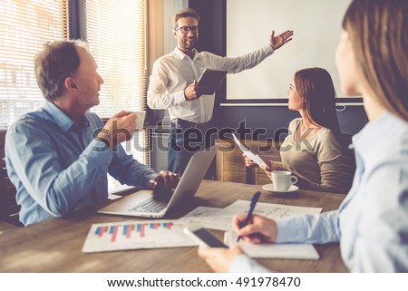 Business people are working in office. Handsome young businessman is making presentation for his colleagues, using a digital tablet and pointing to the board