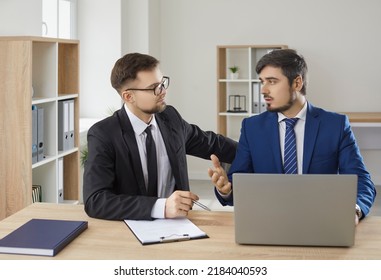 Business People Working Together In Office. Two Serious Men In Suits Sitting At Desk With Laptop Computer And Talking. Team Of Young Lawyers Sharing And Discussing Difficult Cases From Their