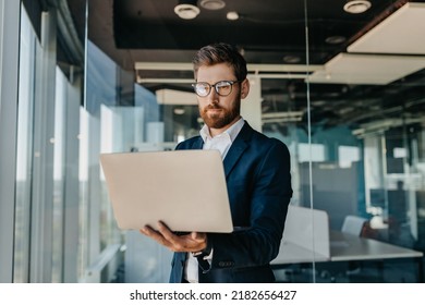 Business person. Successful middle aged businessman with laptop in hands standing in modern office interior. Serious male entrepreneur in formal suit working on computer - Shutterstock ID 2182656427