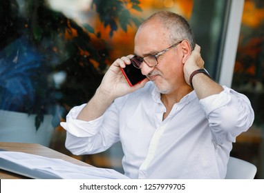 Business Portrait Of An Active And Agitated Mature European Man With A Bald Head And Glasses, Reading A Legal Document While Talking On A Smartphone, Scratching The Back Of His Head From Tension