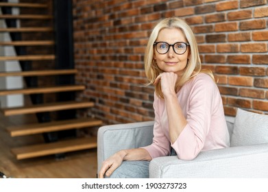 A business portrait of charming middle-aged woman wearing glasses looks at camera, a confident lady sits at armchair at office space, psychologist, consultant. A beautiful friendly blonde indoor - Powered by Shutterstock