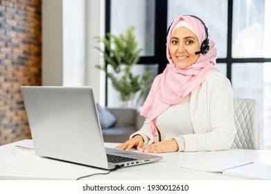 Business portrait of muslim woman wearing hijab, a wireless headset using a laptop for work in the contemporary office. Middle-eastern businesswoman looks at the camera and smiles sitting at the desk