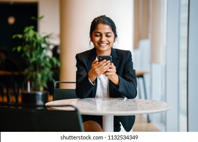 Business Portrait Of A Young Indian Asian Woman Professional In A Suit Sitting At A Desk In Her Office. She Is Smiling As She Is Looking At Her Smartphone.