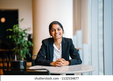 Business Portrait Of A Young Indian Asian Woman Professional In A Suit Sitting At A Desk In Her Office. She Looks, Happy, Hopeful And Confident About Her Future. 