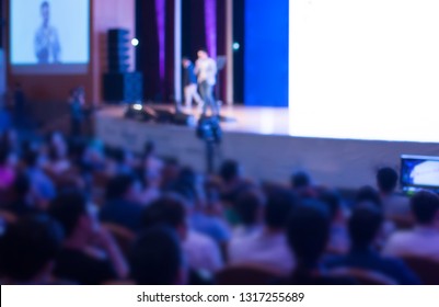 Business Presentation At A Corporate Conference. Blurred Audience Listens To Speaker Giving Public Speech On Stage At Global Executive Symposium.
