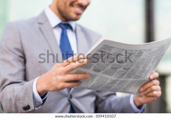business, press, news and people and concept -\
close up of young smiling businessman reading newspaper over office\
building