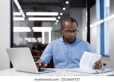 Business professional concentrating on reviewing documents at desk with laptop in modern office. Man appears focused and engaged, highlighting concept of productivity and attention to detail. - Powered by Shutterstock